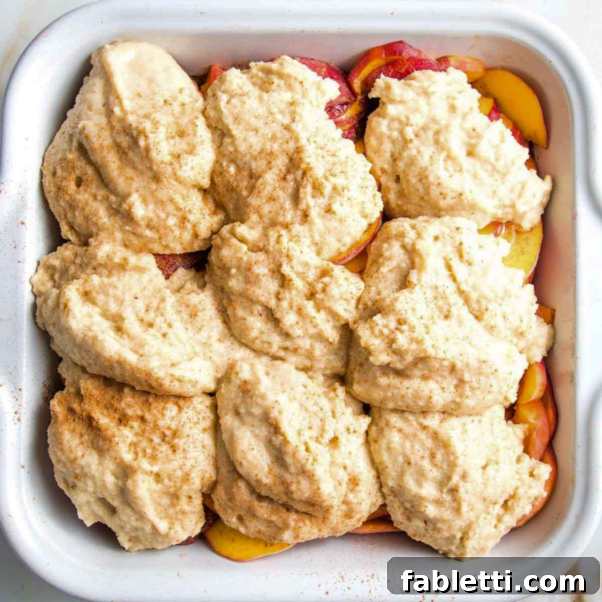 Spoonfuls of cobbler dough scattered over sliced peaches, showing an intentionally rustic appearance.