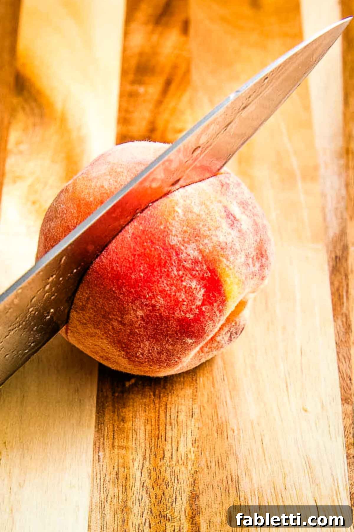 Knife cutting horizontally through the non-stem end of a fresh peach, demonstrating the unique cutting technique.