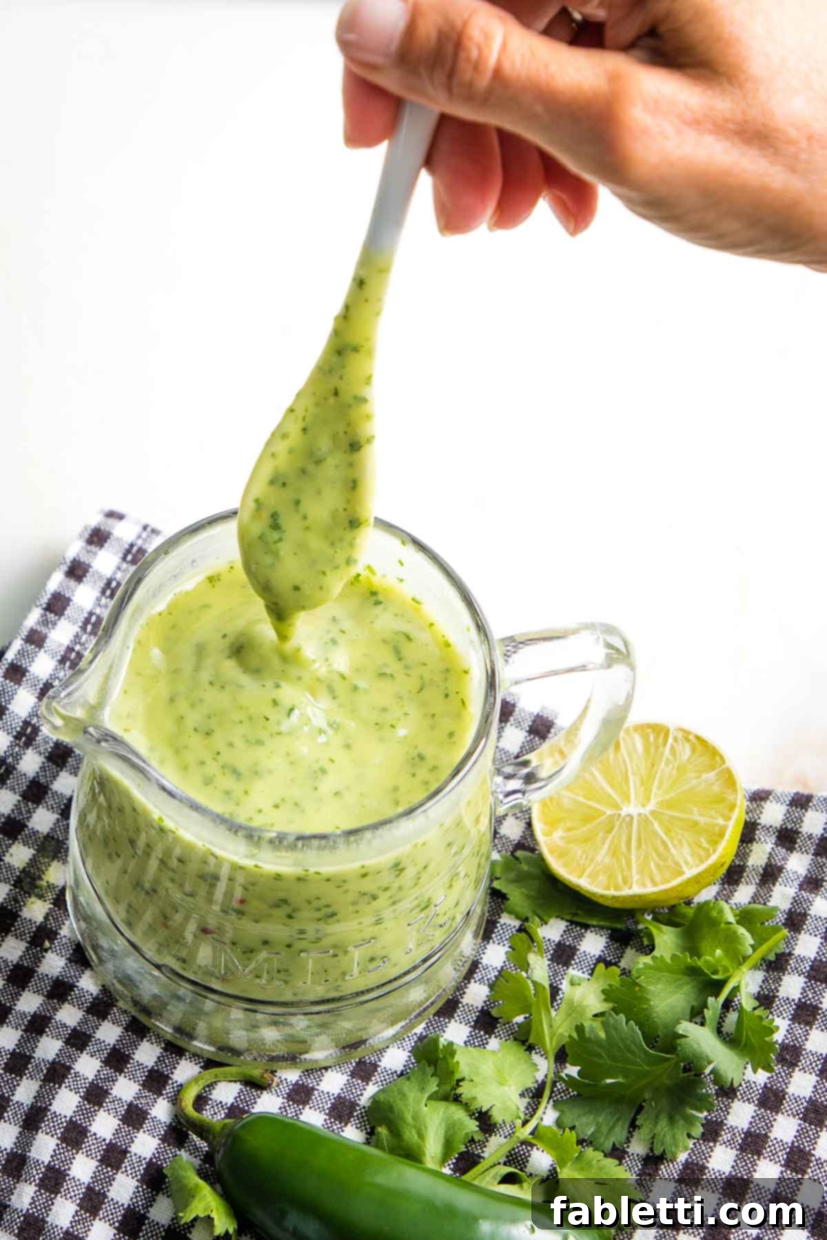 Luscious Dairy-Free Avocado Lime Dressing 2 Light green avocado dressing in a glass pitcher with white spoon, garnished with fresh cilantro and lime, ready to be served.