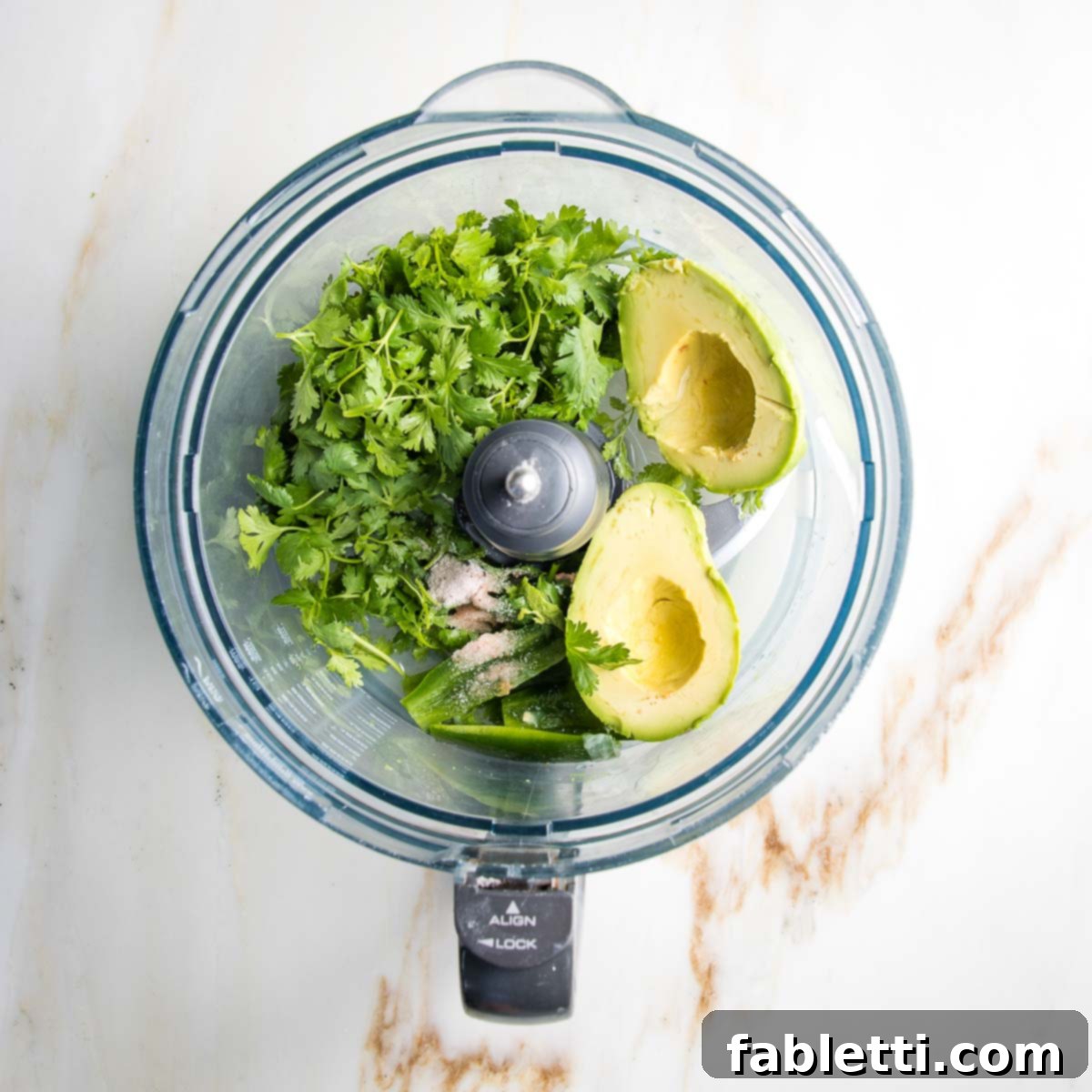 Luscious Dairy-Free Avocado Lime Dressing 4 Food processor bowl containing halved and pitted avocado, fresh cilantro leaves, sliced jalapeno, and a pinch of seasoning, all perfectly arranged and ready for blending into a creamy dressing.