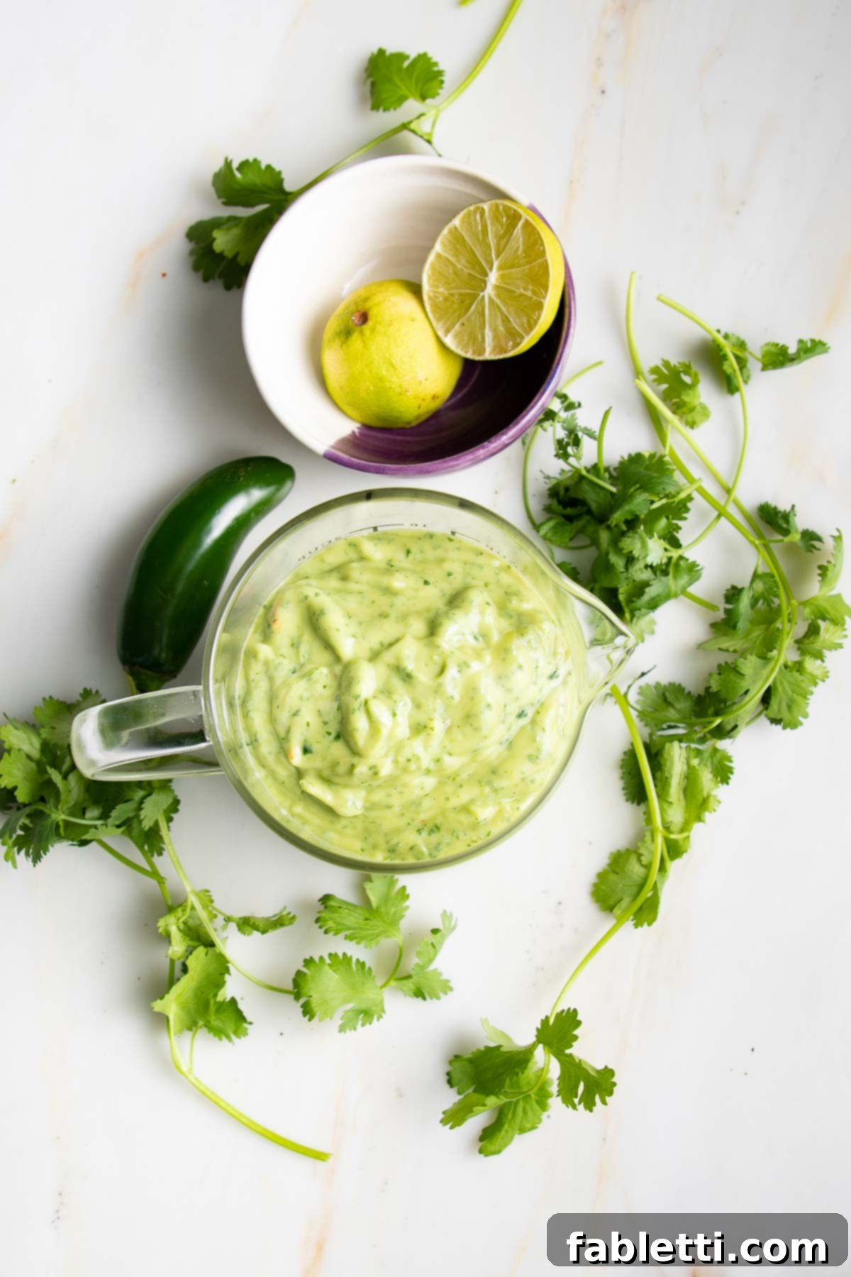 Luscious Dairy-Free Avocado Lime Dressing 6 Avocado dressing in a stylish glass pitcher, garnished with fresh sprigs of cilantro and a vibrant zested lime half, artfully arranged on a clean white surface, ready for pouring.