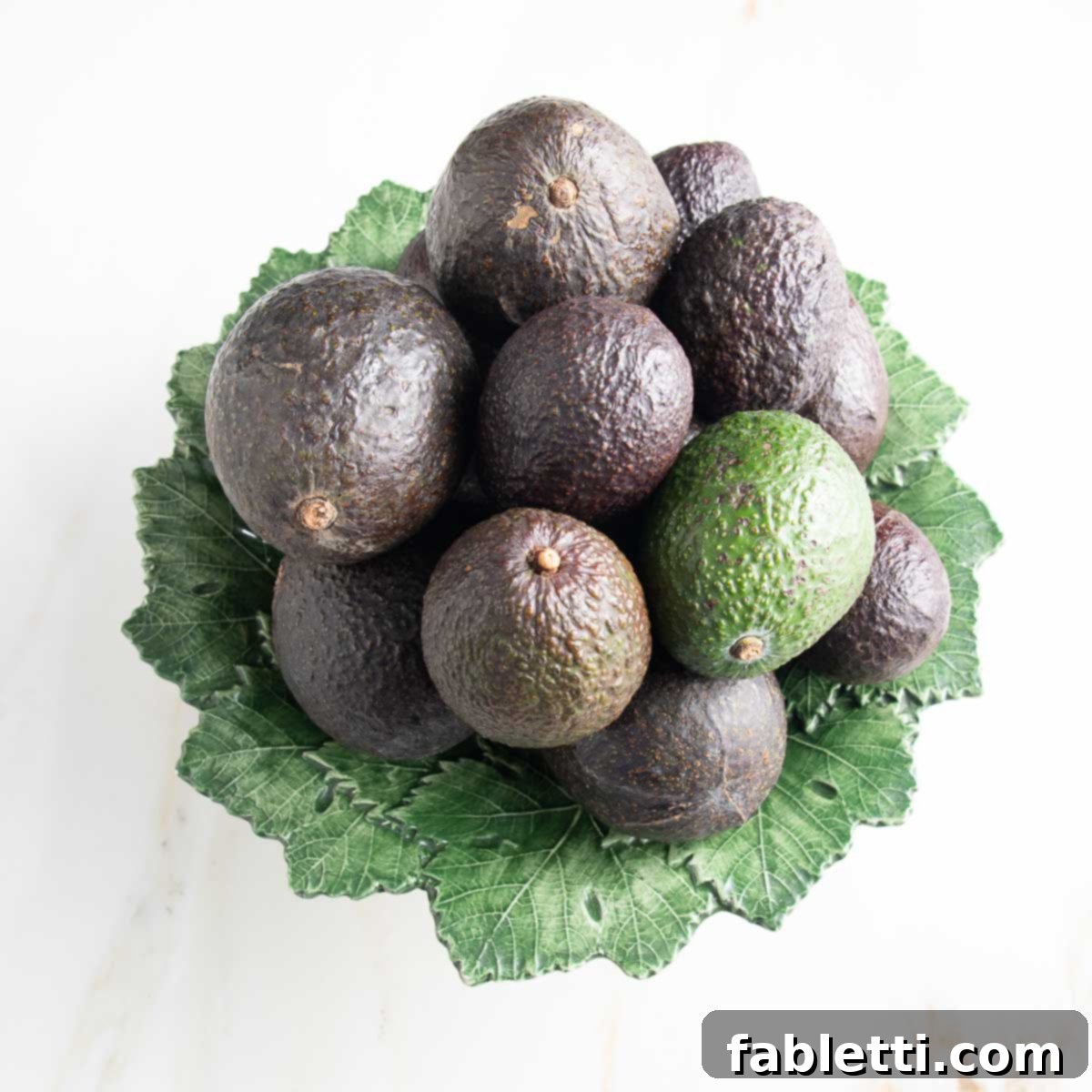 Luscious Dairy-Free Avocado Lime Dressing 7 A vibrant green plate brimming with various whole avocados. One avocado, visibly unripe with a light green skin, stands out among its darker, riper counterparts, illustrating different stages of ripeness.