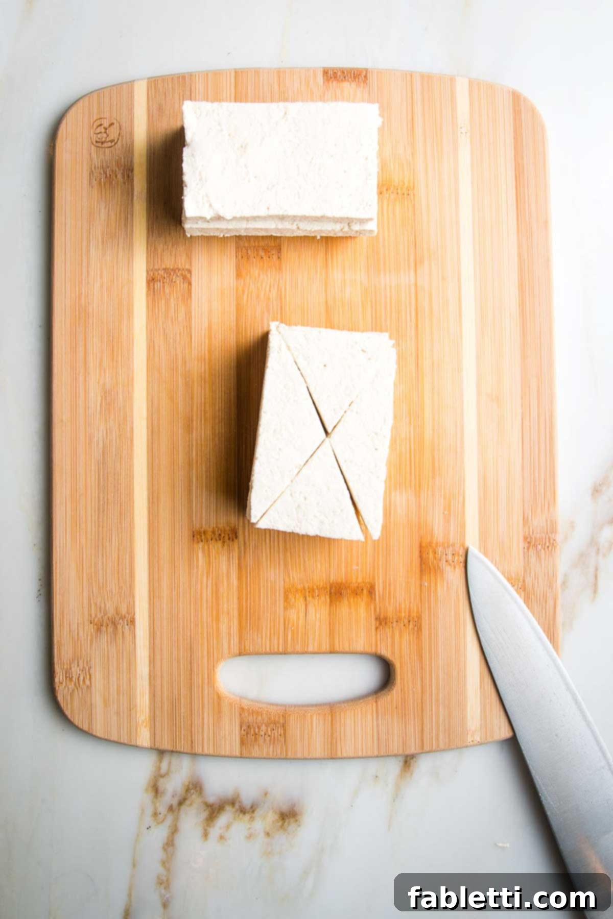 Ultimate Crispy Baked Tofu 6 Slabs of tofu on a wooden board, being cut into bite sized triangular pieces.