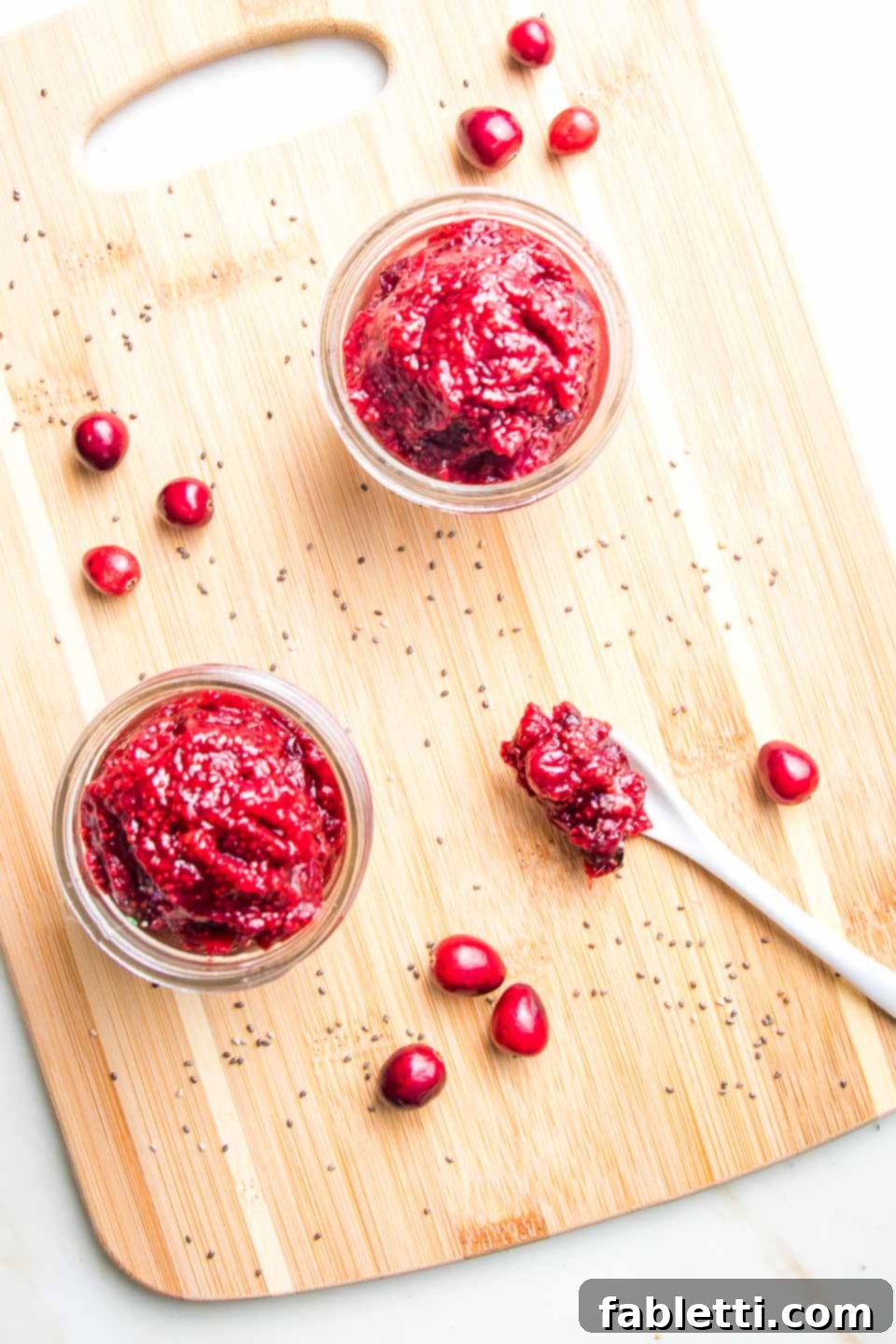 Naturally Sweetened Cranberry Chia Jam 8 Small jar of cranberry chia jam on a wooden board with a few fresh cranberries and some chia seeds sprinkled about.