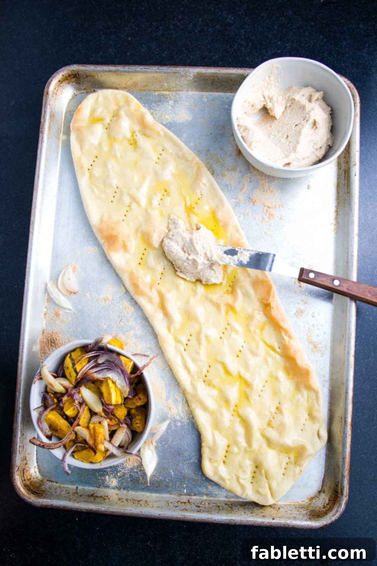 Effortless Plant-Based Flatbread 13 Spreading hummus onto a flatbread pizza crust, brushed with oil.