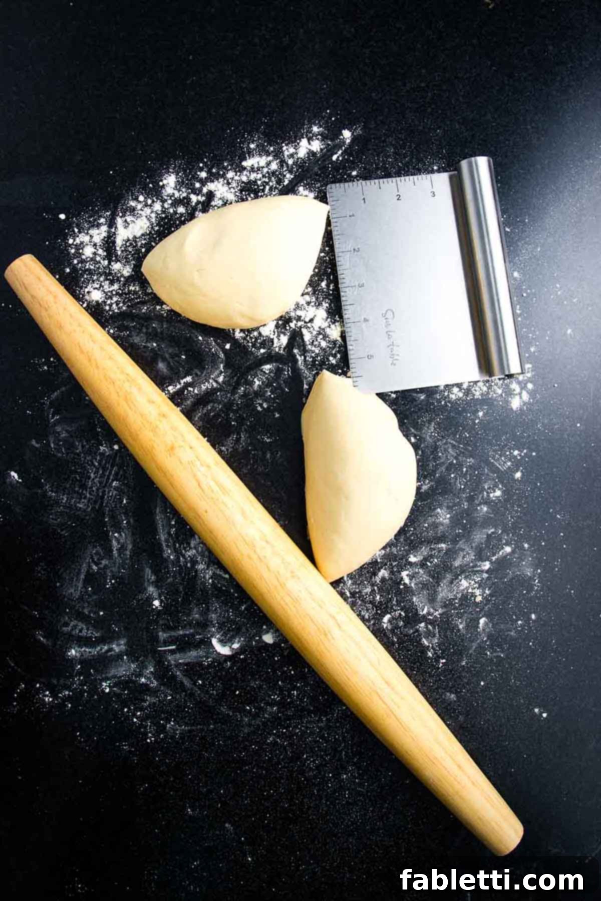 Effortless Plant-Based Flatbread 7 Ball of pizza dough cut in half on a floured black surface with wooden rolling pin off to the side.