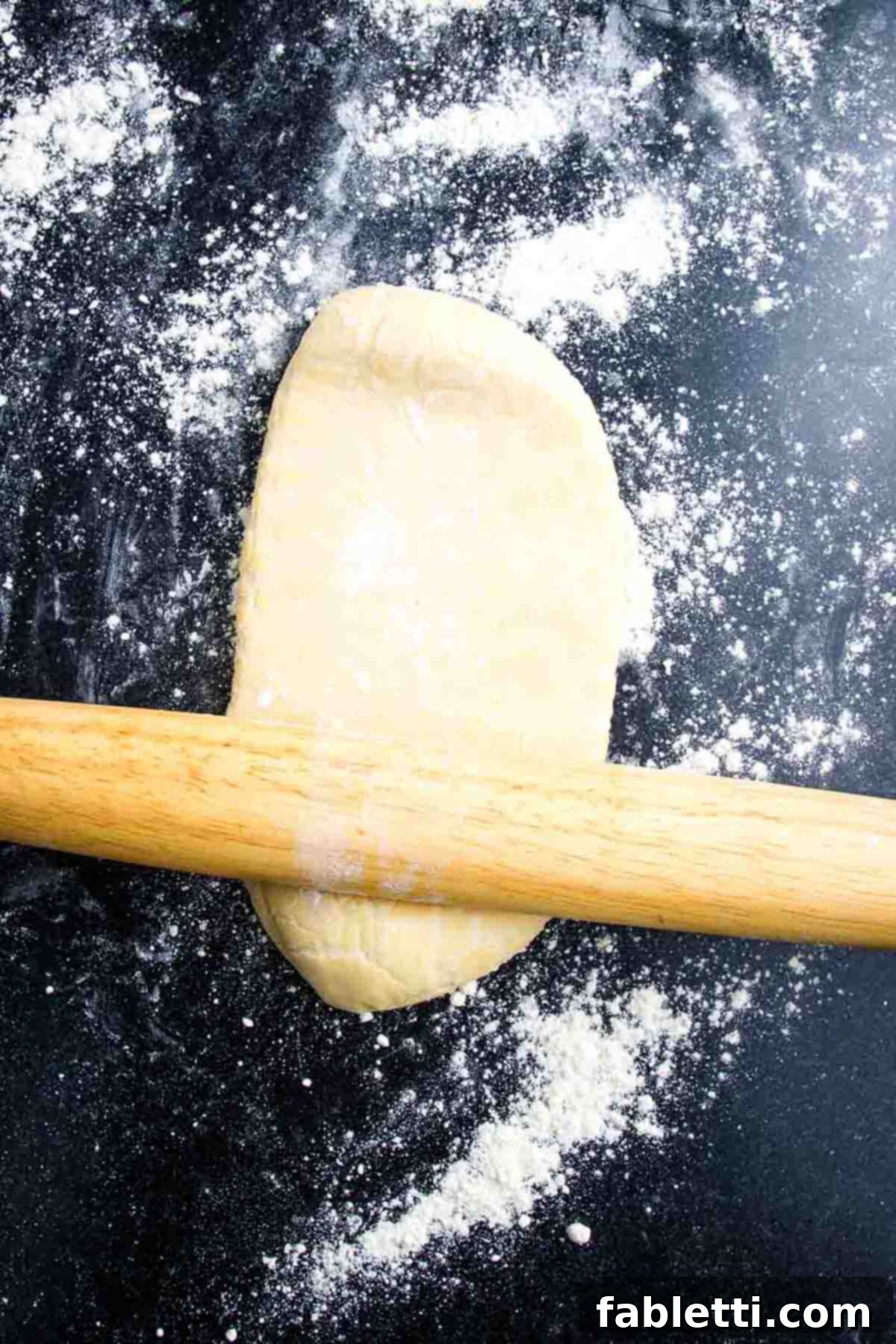 Effortless Plant-Based Flatbread 8 Using a wooden rolling pin to roll out pizza dough on a black surface sprinkled with flour.