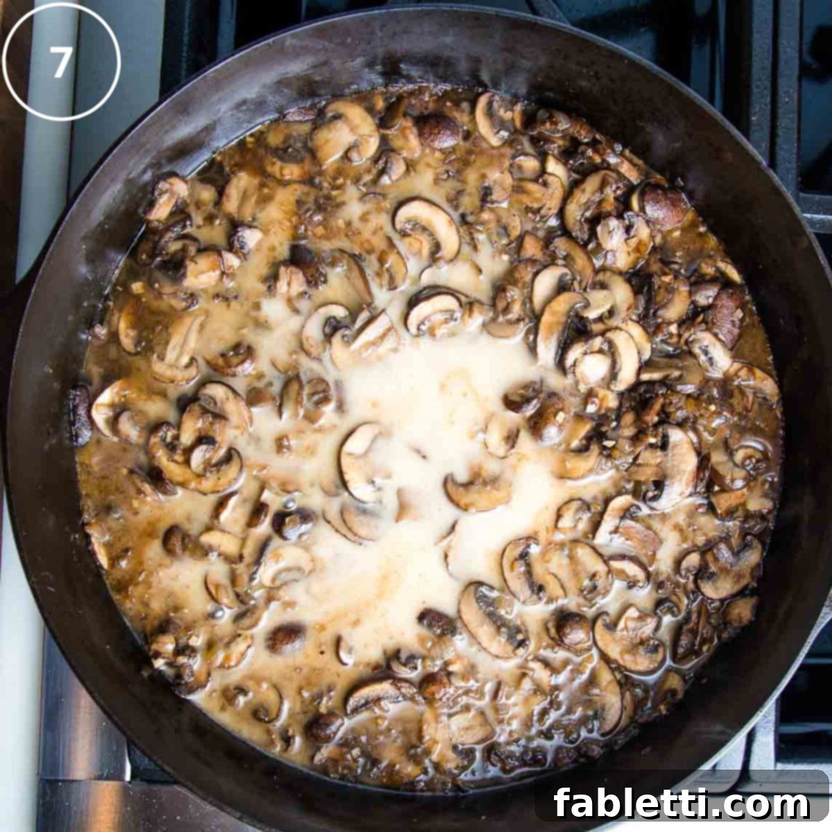 Hearty Vegan Mushroom Stroganoff 11 A splash of white liquid (arrowroot slurry) being stirred into the rich brown mushroom sauce in a cast iron skillet, indicating the thickening stage.