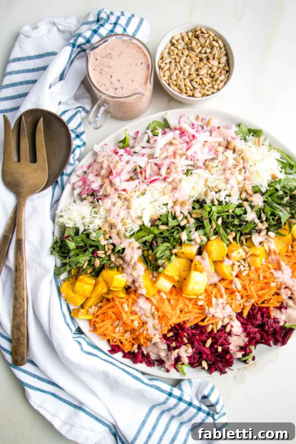 A beautifully arranged Shredded Rainbow Salad featuring radishes, green and purple cabbage, vibrant beet greens, carrots, red beets, and golden roasted delicata squash cubes on a pristine white rimmed plate. A small bowl of pink, creamy dressing and a separate bowl of sunflower seeds are also present, highlighting the salad's fresh ingredients and enticing presentation.
