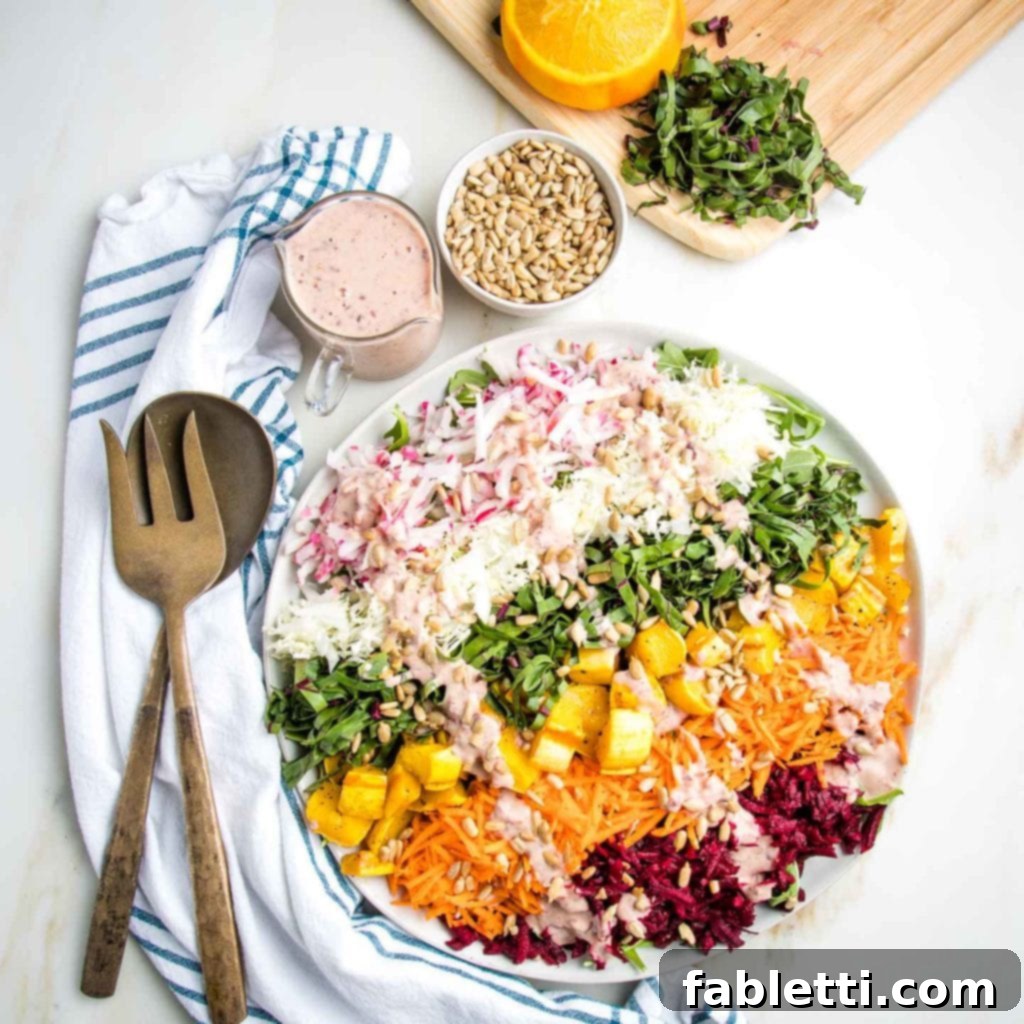 A vibrant Shredded Rainbow Salad elegantly arranged on a white plate, featuring distinct colorful stripes generously drizzled with a creamy pink dressing. The setting includes serving utensils on a white tea towel with blue stripes, a small pitcher of extra dressing, a bowl of sunflower seeds, and in the background, a cutting board with julienned beet greens and squeezed oranges, emphasizing freshness and readiness for serving.