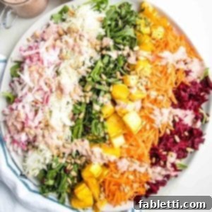 Shredded rainbow salad on a round rimmed white plate: a layer of arugula covered with shredded radish, green cabbage, beet leaves, roasted delicata squash, shredded carrots, purple cabbage and red beets.