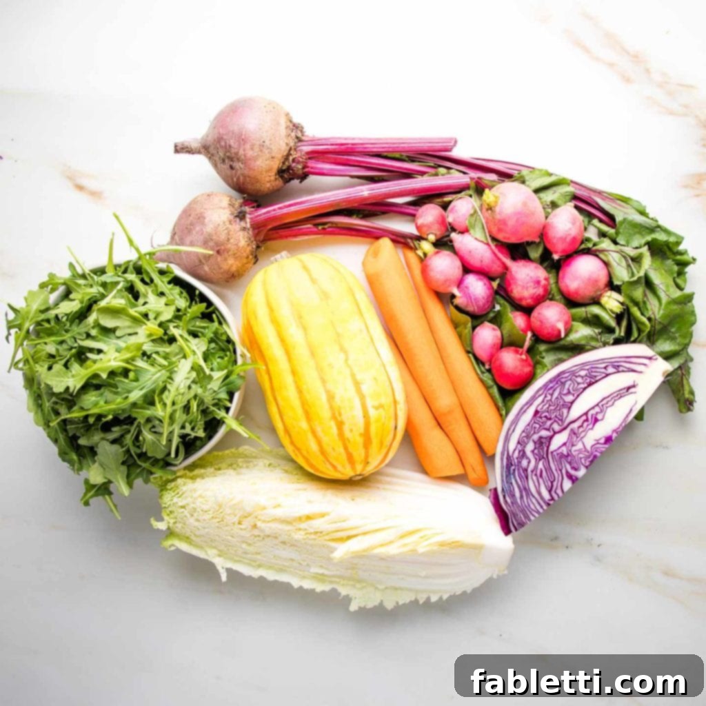 A pre-chopped assortment of vibrant ingredients for a rainbow salad laid out on a clean surface. The selection includes a small bowl of fresh arugula, a quarter of green cabbage, a quarter of purple cabbage, a whole delicata squash, two red beets with their leafy greens still attached, a bunch of bright radishes, and three peeled carrots, ready for preparation.