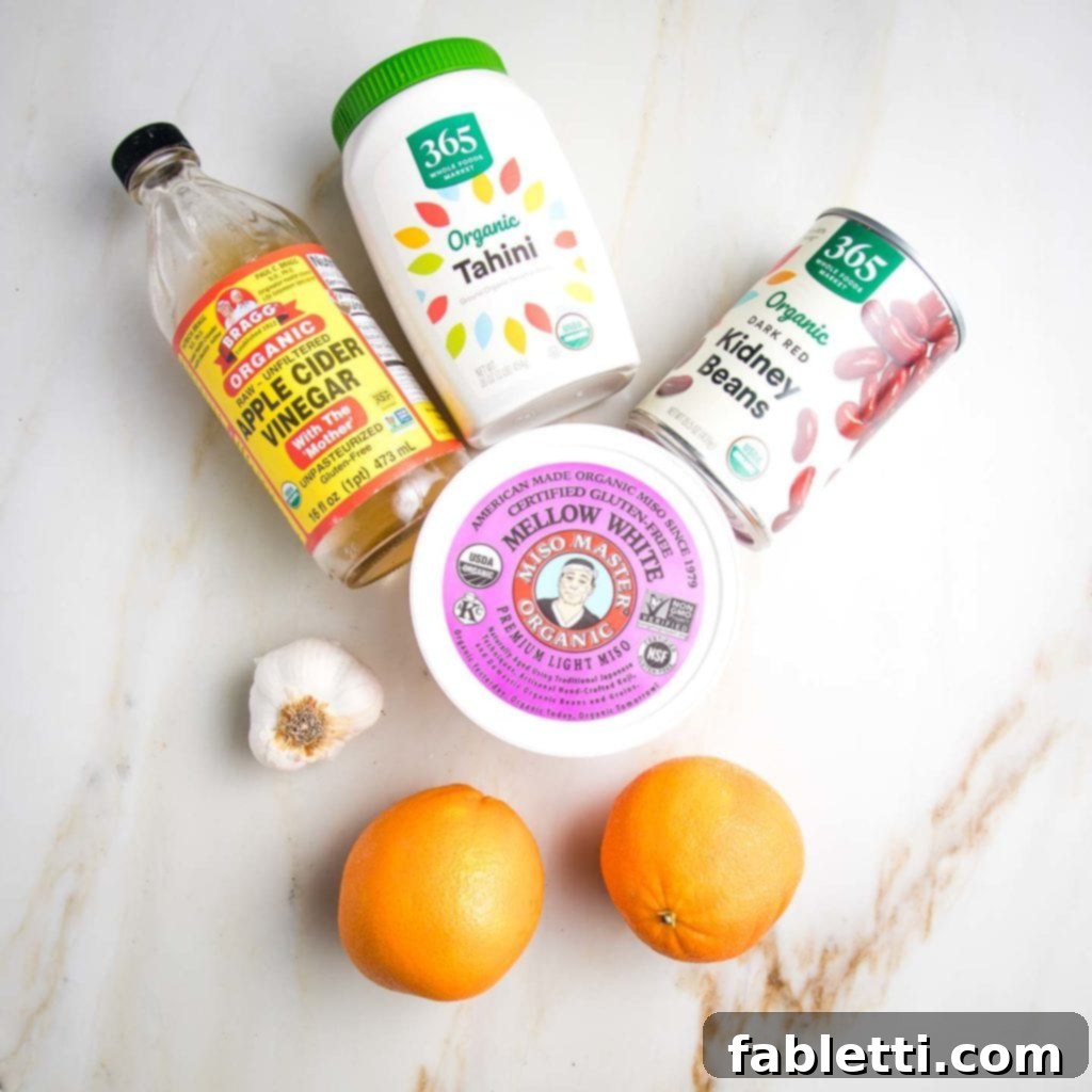 Ingredients for a creamy, no-oil salad dressing arranged on a marble countertop. The components include a bottle of apple cider vinegar, a head of fresh garlic, two whole oranges, a jar of tahini, a can of red kidney beans, and a container of light miso paste, showcasing the wholesome and flavorful elements of the dressing.
