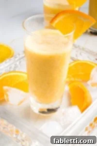 Frothy Orange Julius Delight 11 Glass filled with a frothy Orange Smoothie, with a wedge of orange on the side of the glass, more orange wedges on the glass serving tray along with some ice cubes.