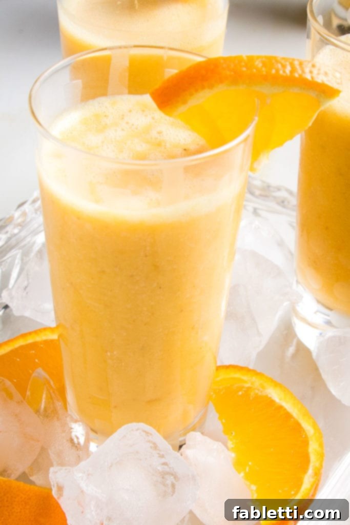 Frothy Orange Julius Delight 3 A close-up of a glass filled with an orange smoothie, garnished with an orange wedge. Ice cubes and orange slices are scattered on a tray next to it, enhancing the vibrant display.