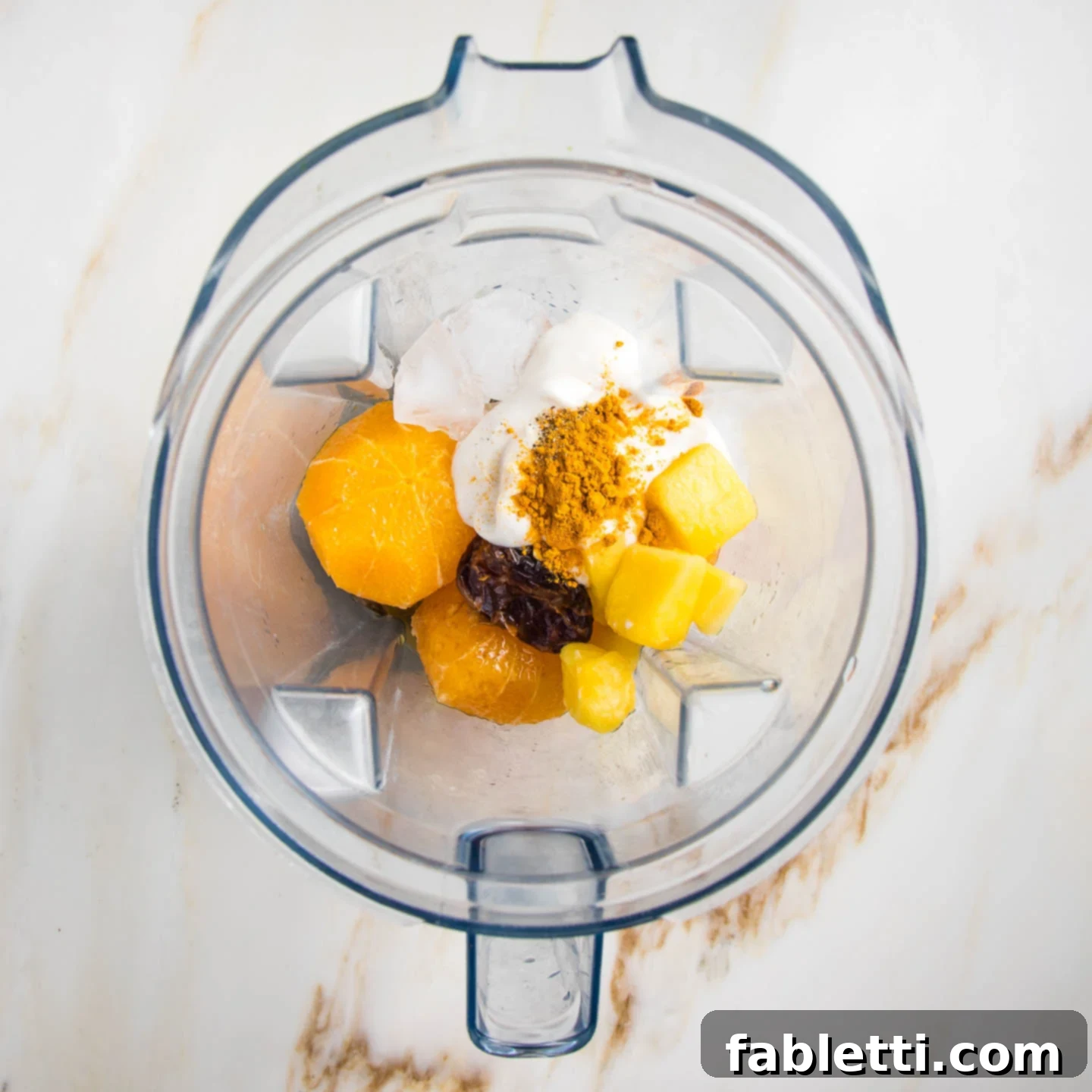 Frothy Orange Julius Delight 6 A blender pitcher filled with the main ingredients for an orange smoothie, including peeled oranges, pineapple chunks, plant-based yogurt, a date, and turmeric powder, ready to be blended.
