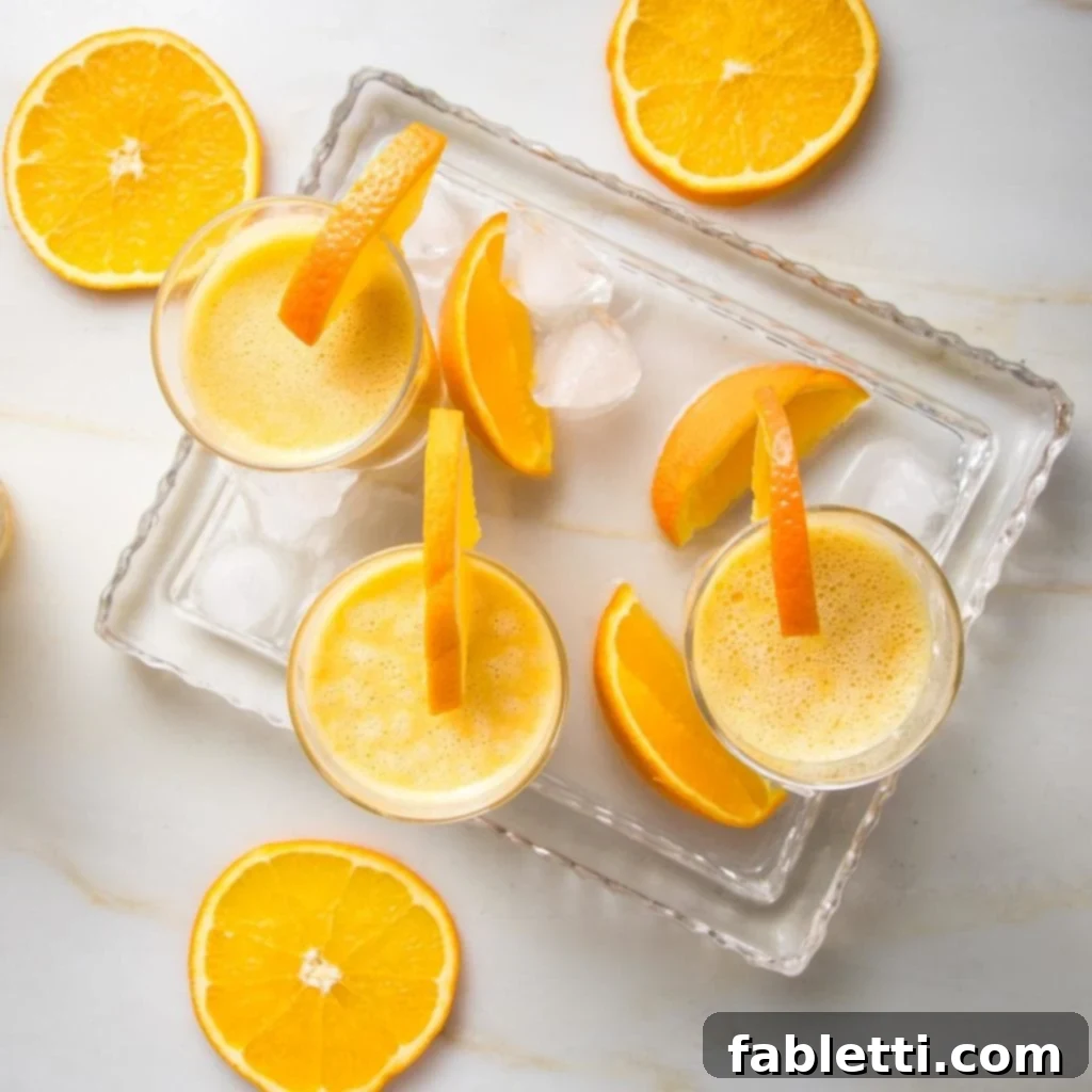 Frothy Orange Julius Delight 9 A glass serving tray holds three frothy Orange Julius smoothies, each garnished with a fresh orange wedge. Ice cubes and more orange slices are scattered around the glasses on the tray and countertop.