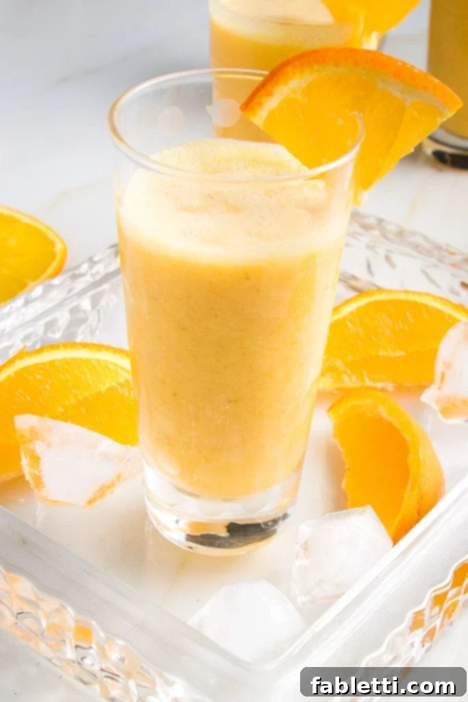 Frothy Orange Julius Delight 10 A single glass of frothy Orange Smoothie, garnished with an orange wedge, resting on a glass serving tray with additional orange wedges and ice cubes scattered around.
