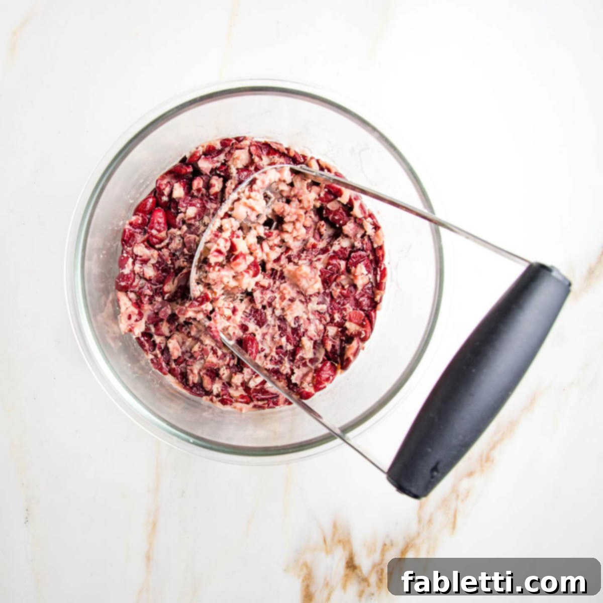 A potato masher expertly squishing vibrant red kidney beans in a clear glass bowl, preparing them for the veggie burger mixture.