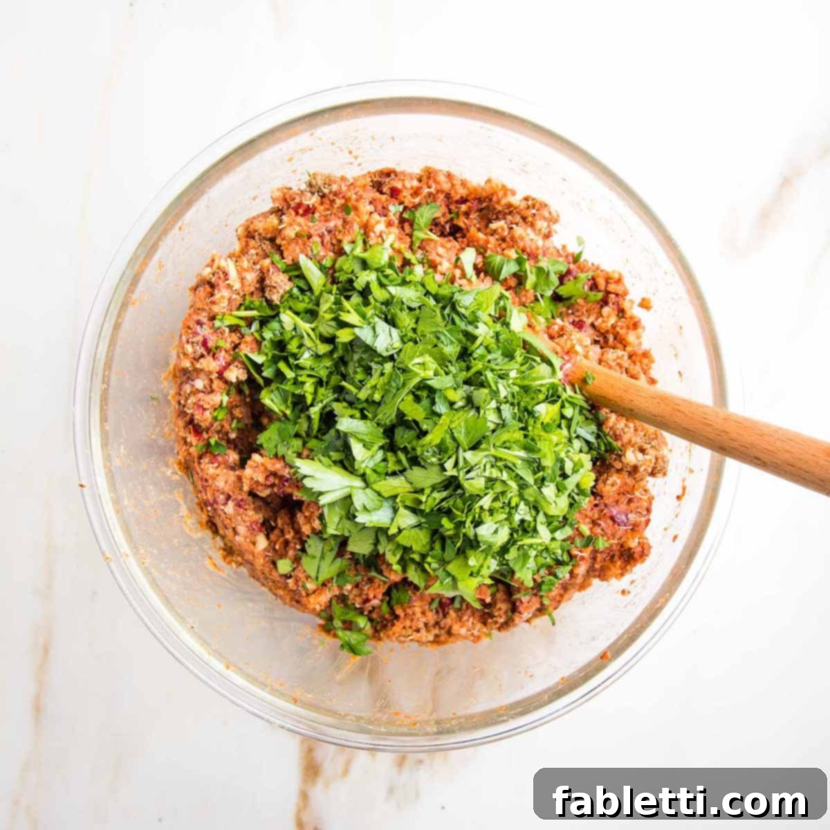 Vibrant finely chopped fresh parsley generously sprinkled over the reddish-brown veggie burger mixture in a glass bowl, adding a final touch of color and aroma.