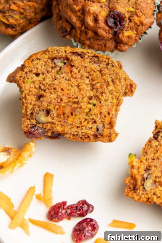 A breakfast muffin expertly cut in half, revealing its rich interior packed with shredded carrots, zucchini, crunchy walnuts, and vibrant dried cranberries. A few cranberries and shredded carrots are artfully arranged on a white plate in the foreground, ready to be enjoyed.