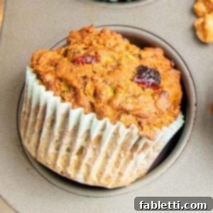 A close-up of a Morning Glory Muffin, featuring a visible dried cranberry and shredded carrots. The muffin, still in its light blue paper liner, is resting outside the baking pan.