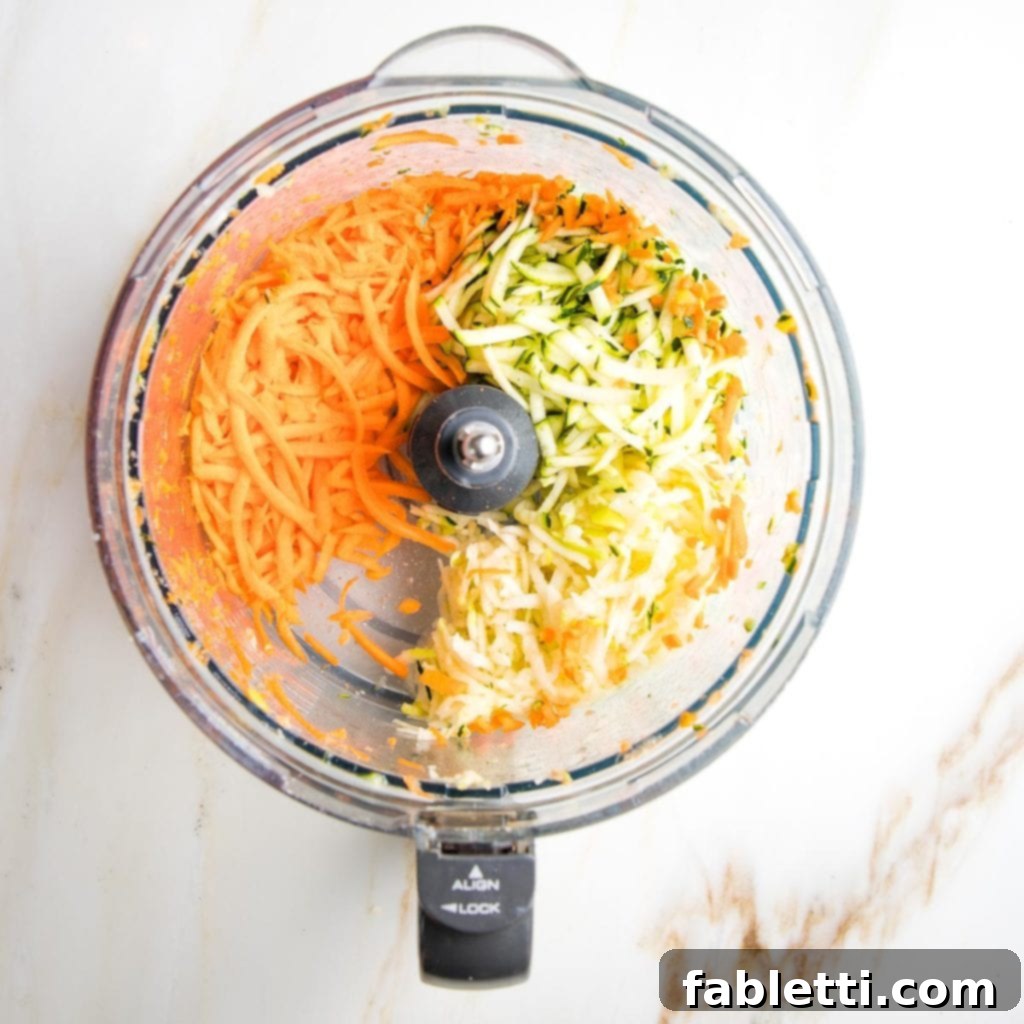 Ingredients for Morning Glory Muffins, including shredded carrots, zucchini, and apple, laid out on a cutting board next to a food processor and a box grater, ready for preparation.