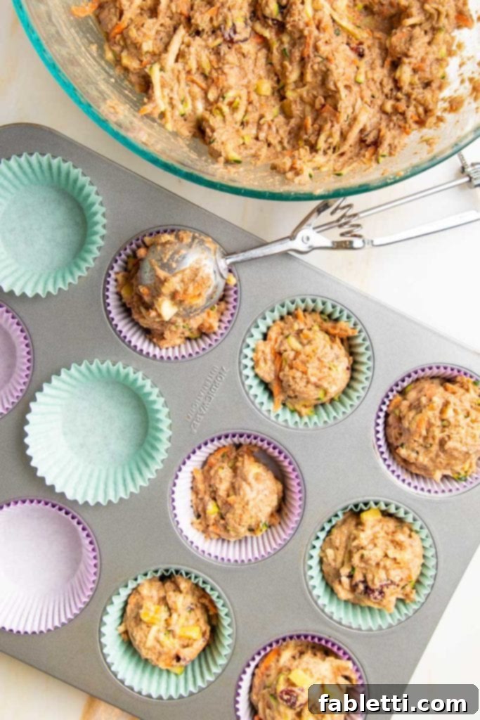 A bowl of Morning Glory Muffin batter sits beside a muffin tin lined with colorful purple and light blue liners. Half of the liners are already filled with batter, and a large cookie scoop is shown filling another, illustrating the easy portioning process.