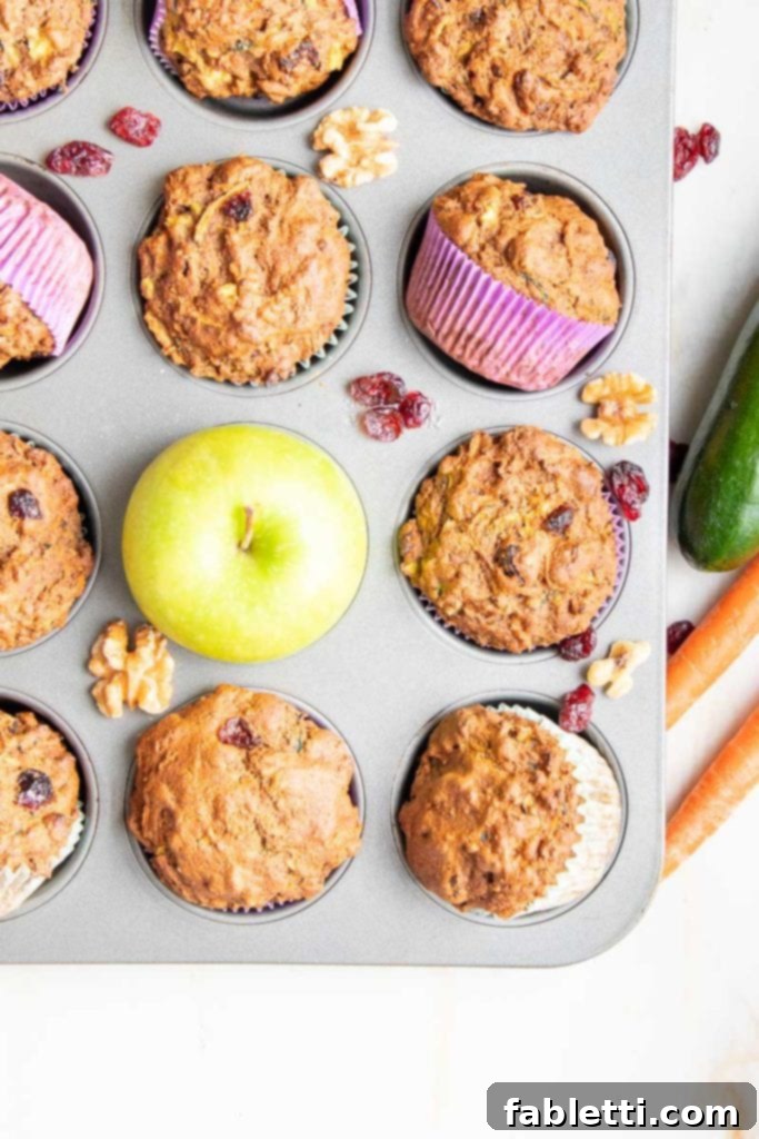 A muffin tin overflowing with freshly baked Morning Glory Muffins, accompanied by a vibrant green apple, scattered dried cranberries and walnuts, and fresh zucchini and carrots on the side, highlighting the natural ingredients.