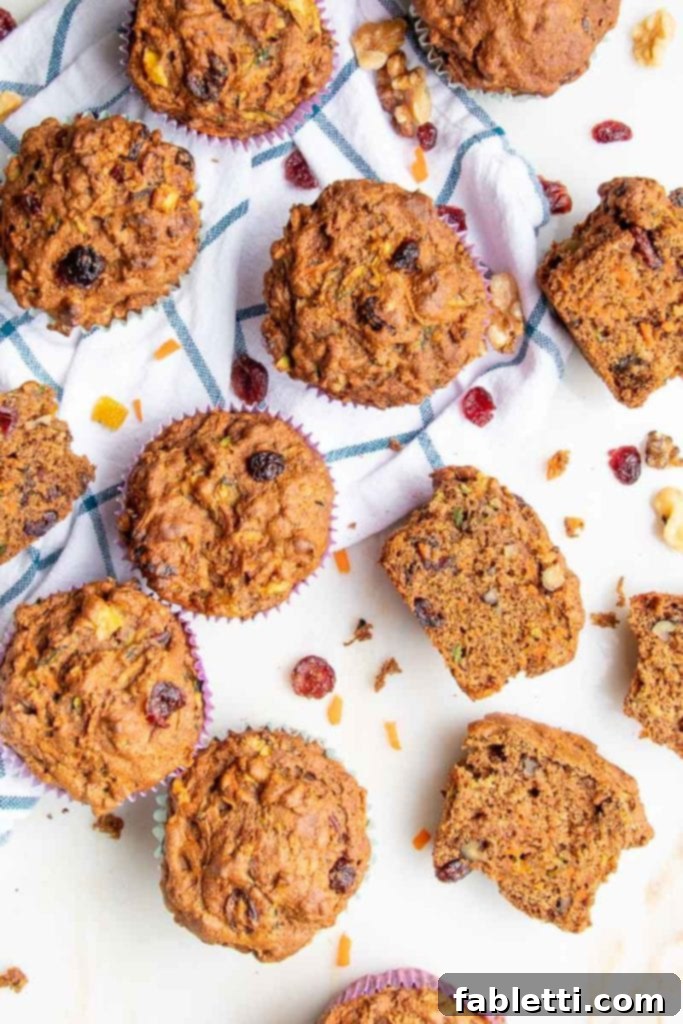 A collection of Morning Glory Muffins scattered on a blue wide plaid tea towel. Some muffins are cut open to reveal their wholesome interior, rich with walnuts, shredded carrots, zucchini, and dried cranberries, showcasing their hearty texture.
