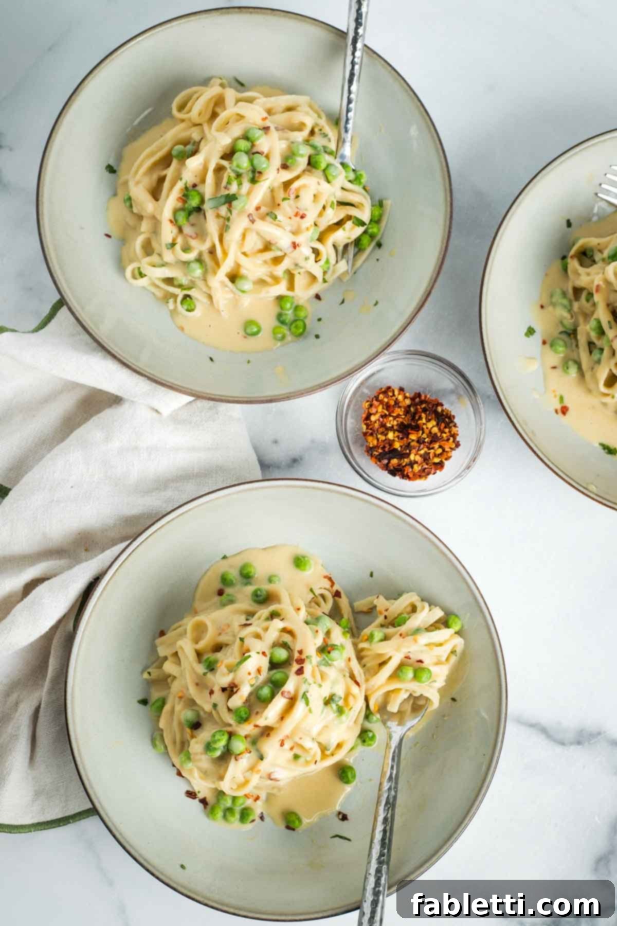 Bowls of creamy vegan Fettuccine Alfredo with green peas, garnished with fresh herbs and a sprinkle of crushed red pepper flakes.