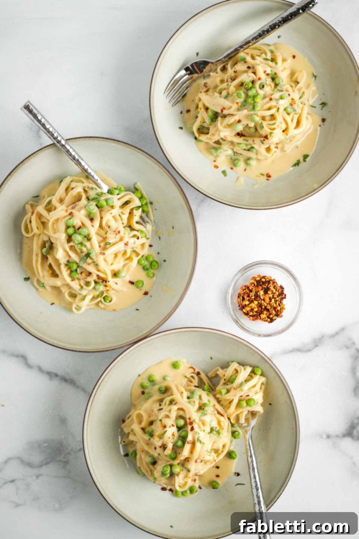 Bowls of creamy vegan Fettuccine Alfredo with peas, crushed red pepper flakes, and fresh herbs.