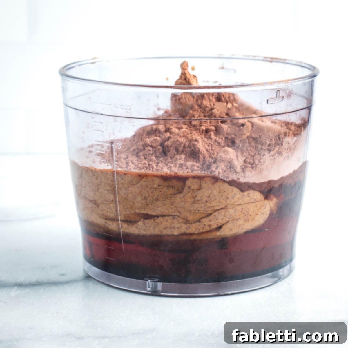 A clear view of a food processor bowl containing almond butter, maple syrup, and cacao powder, ready to be blended.