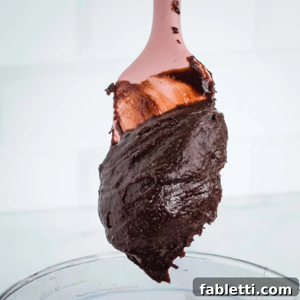 A thick, luscious chocolate frosting clinging firmly to a pink spatula, showcasing its rich consistency.